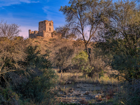 A weathered castle overlooks a tranquil scene filled with lush trees and a gentle stream. The setting sun casts a warm glow, creating a peaceful atmosphere in nature.の写真素材