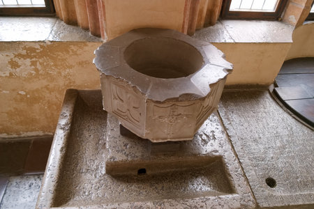 Inside an old church, a stone baptismal font sits prominently. Its detailed carvings reflect the craftsmanship of a bygone era. Sunlight filters through nearby windows, illuminating the scene.の写真素材