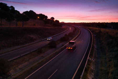Cars and trucks drive along a winding highway as the sun sets, casting colorful hues in the sky. Trees line the road on a tranquil evening.の写真素材