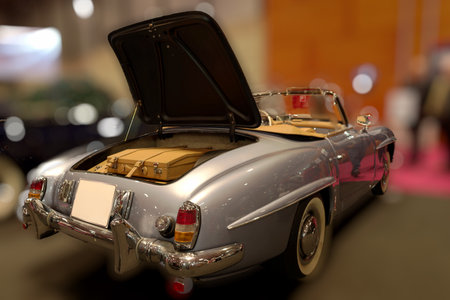 A classic convertible car is showcased indoors at a vintage automobile show. The trunk is open, revealing stylish luggage, while the event buzzes with enthusiasts admiring the vehicles.の写真素材