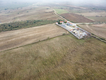 Aerial view shows a storage facility located near open fields and empty land. The landscape is mostly flat with a few trees and a distant road. Cloudy weather covers the scene.の写真素材