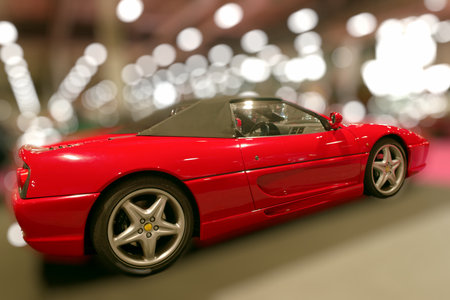 A red sports car is parked at an indoor auto show. Bright lights shine in the background creating a...