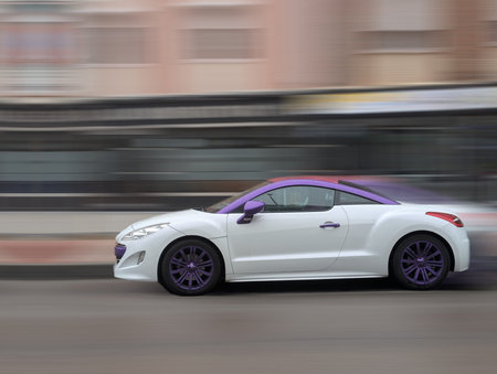 A white car with purple accents is speeding down a city street during the day. The scene captures...