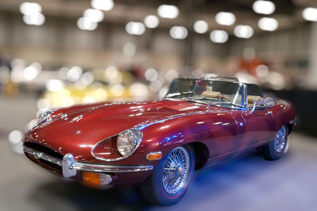 A red convertible car is showcased in a large exhibition hall. People walk around, admiring the...