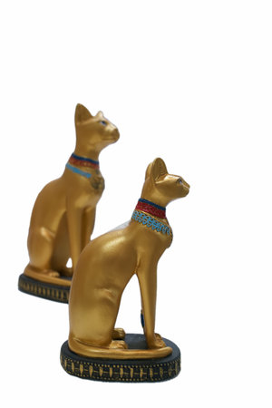 Two golden cat figurines are sitting upright facing forward. They are decorated with colorful collars and rest on a dark base. The background is plain and bright.の写真素材