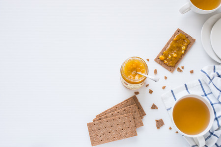 Brown rye crispy bread (Swedish crackers) with orange jam and pieces of orange, with cups filled with herbal tea, on white background with space for text, top view. Meal conceptの写真素材