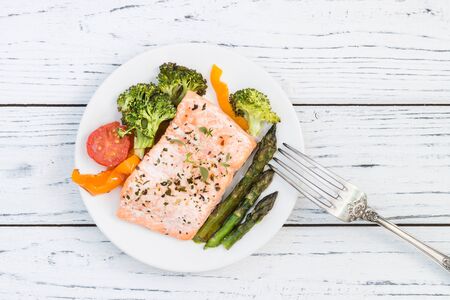 Baked salmon fillet with pepper, red cherry tomatoes, asparagus, broccoli and fresh thyme served on white plate. Wooden table. Lunch or dinner concept. Top viewの写真素材