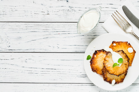 Fried pineapple slices with creme fraiche, mint in white plate on wooden background. Top view, copy spaceの写真素材