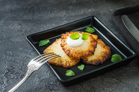 Fried pineapple slices with creme fraiche, mint in black square plate on dark backgroundの写真素材