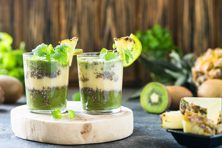 Healthy layered smoothie with kiwi, pineapple, chia seeds, mint. Two glasses on wooden board, dark backgroundの写真素材