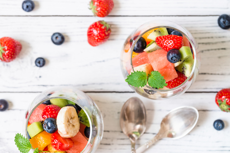 Healthy fresh fruit salad in transparent glasses with spoons on wooden table. Top view. Delicious summer meal: strawberries, blueberries, oranges, kiwi, banana, watermelon, balm mintの写真素材
