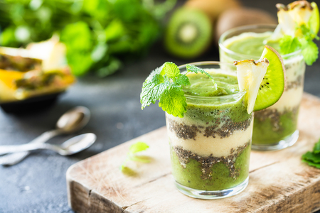 Healthy layered smoothie with kiwi, pineapple, chia seeds, mint. Two glasses on wooden board, dark backgroundの写真素材