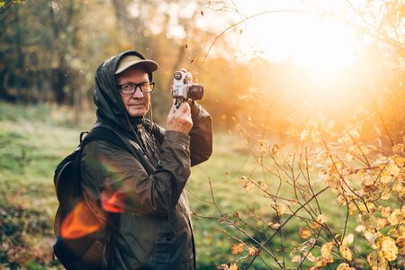 Handsome senior man photographer standing in autumn park and taking pictures with camera.