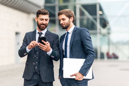 Young businessman in suit using mobile phone and showing something to his colleague while they standing near the modern building outdoors.の写真素材