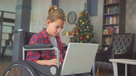 teenage girl disabled in a wheelchair, sitting at the laptop, against the Christmas treeの写真素材