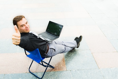 Image of the businessman sitting with the laptop and looking at the camera in the empty street. Focus is made on top of the background marbled tile in the street.の写真素材