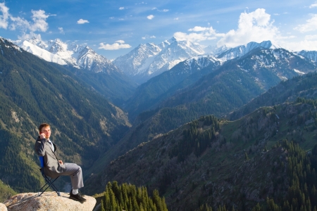 Image of a young businessman who sits on a chair at the top of the mountain and looking at the camera, talking about new idea by phone.の写真素材
