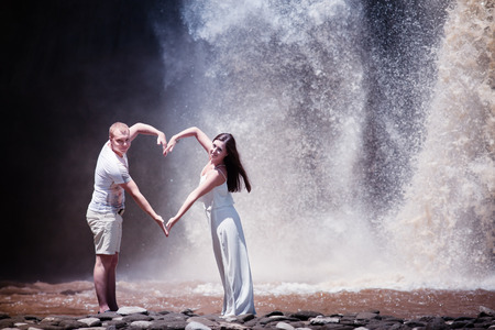Photo of young couple with love sign by hands, near the waterfallの写真素材