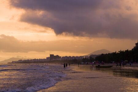 Image of sunset on the beach in Hainan. Many people walking through coastline and swimming on the background.の写真素材