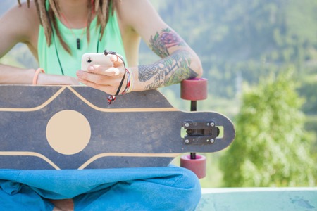 Closeup of hippie young and handsome man with skateboard outdoor at mountain. He using mobile phone and listening to musicの写真素材