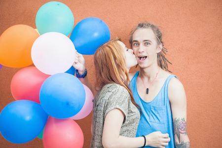 Young happy couple near the orange wall stand with balloons. The girl kisses the young man's cheek. Man smiling enthusiasticallyの写真素材