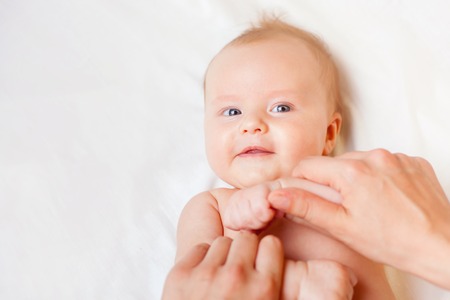 Mother makes massage and stretch for baby hands, with happy newborn at white bed, apply oil on the handの写真素材