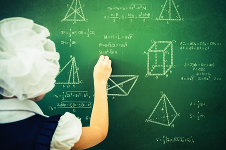 Smart student or schoolgirl drawing mathematic formula at blackboard, dressed in school uniform, scribbled with chalk - formulas and drawing. Concept of intelligent and scientist girlの写真素材