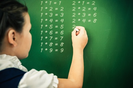 First grade happy schoolgirl wrote on blackboard multiplication table with chalk at classroom. Little girl dressed in a school uniform. Concept of knowledge day and first of septemberの写真素材