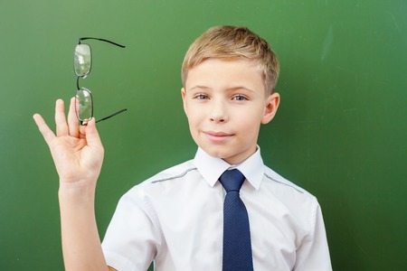 Successful schoolboy standing near the blackboard in a school classroom, dressed in a school uniform, and holding medical glasses. Boy's poor eyesightの写真素材
