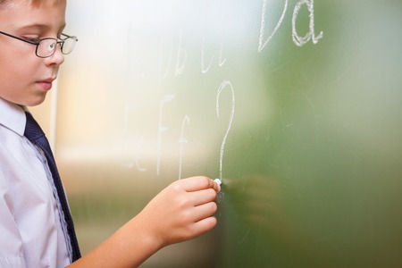 School boy writes English alphabet with chalk on a blackboard in school classroom. He does not know...