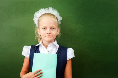 First grade schoolgirl holding a notebook at classroom. Little girl dressed in a school uniform. Concept of knowledge day and first of september. There is a copy space at green blackboardの写真素材