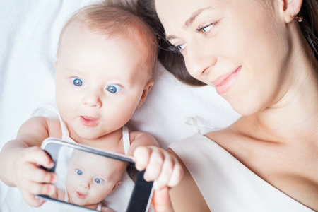 Funny baby girl make selfie on mobile phone and lying near her mother on a white bed. Newborn...