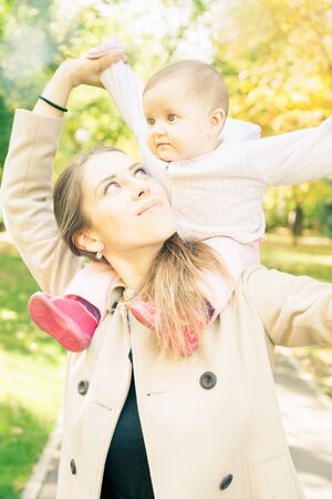 Beautiful mother with her baby daughter outdoor at autumn park. Concept of happy family. Child sitting on her mom's neckの写真素材