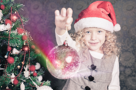 Cute girl decorating a christmas tree at home. Holding magical red decoration ball. Holiday concept, New Year, giftの写真素材