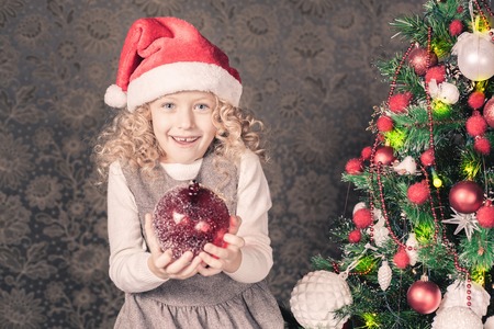 Cute girl decorating a christmas tree at home. Holding magical red decoration ball. Holiday concept, New Year, giftの写真素材