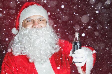 Happy Santa Claus holding Cola fresh beverage at glass bottle. Christmas holiday, New Year. Snowy winter blizzard with magical snow and snowflakes at backgroundの写真素材