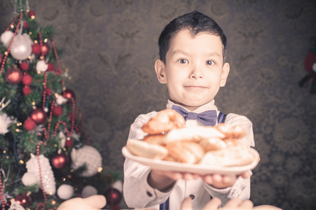 Handsome asian boy giving to Santa Claus cookies at Christmas holiday. Plate of biscuits, sweet baking. Decorated Christmas tree at living roomの写真素材