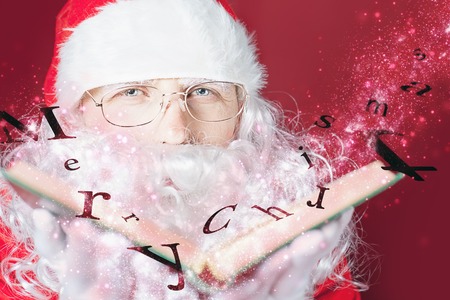 Santa Claus reading a book with Christmas fairy tale. Mature man blowing on the letters of Merry Christmas that fly with magical glitter and stardust.の写真素材