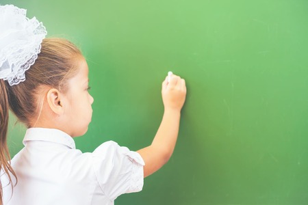 First grade happy schoolgirl wrote on blackboard with chalk at classroom. Little girl dressed in a school uniform. Concept of knowledge day and first of septemberの写真素材