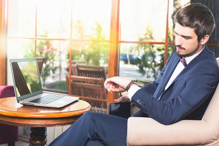 Handsome man looking time at watch. Waiting and sitting at cafe or restaurant. Businessman dressed in fashion costume. Stylish man. Laptopの写真素材