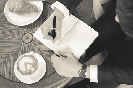 Close-up image of coffee break with a cap of coffee and croissant. Black and white, retro. Businessman writes something in notebook, office. Good Morning monday. Cafe, restaurantの写真素材