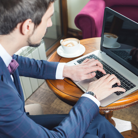 Young businessman working at laptop, typing text or message at social network. Coffee break. A cup of coffee at the table. Internet using wi-fi connectionの写真素材