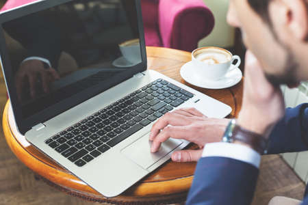 Young businessman working at laptop, typing text or message at social network. Coffee break. A cup of coffee at the table. Internet using wi-fi connectionの写真素材