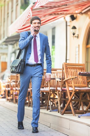 Successful businessman talking by mobile phone and walking at street near french cafe. Early morning. Urban, city life. Happy intelligent man.の写真素材