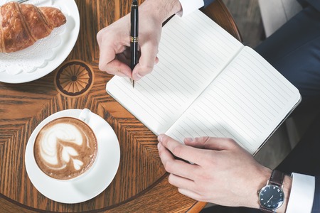 Close-up image of coffee break with a cap of coffee and croissant. Businessman writes something in notebook, office. Good Morning. Cafe, restaurantの写真素材