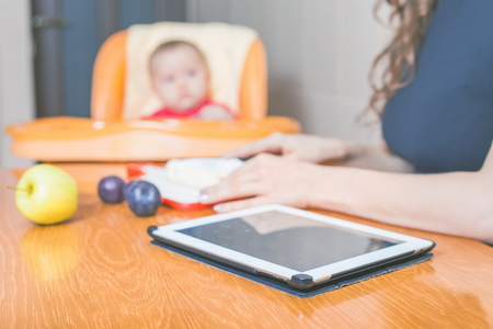 Mother is looking for a recipe of preparation of baby food. Tablet pc computer, internet, wifi. Kitchen, home. Fresh fruit, banana, apple, prunes are on the tableの写真素材