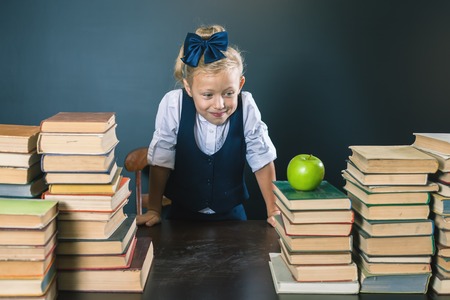 Cute school girl standing near the table with many books and looking at one green apple. Healthy...