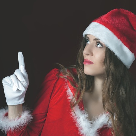 Attractive sexy woman dressed in Santa hat pointing at copy space. Brunette babe. Christmas night...