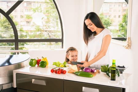 Cute little girl cooking with her mother in the bright modern kitchen. Healthy food, cooking healthy salad with vegetables ingredients. Mom and daughter looking the recipe on mobile phoneの写真素材