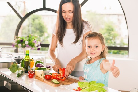 Cute little girl cooking with her mother and shows thumbs up. Healthy food, cooking healthy salad with vegetables ingredients. Mom and daughter cooking together. Recipe food for baby or childの写真素材